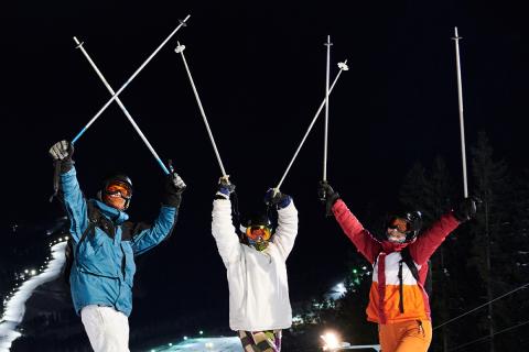 Three skiers posing, holding their poles up in the air on top of a mountain at night.