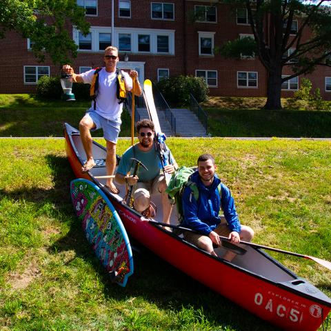 Students outside posing with a canoe, life jackets, a surfboard, skis and other outdoor gear
